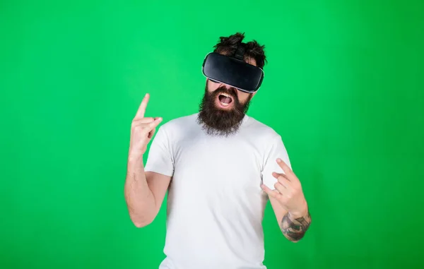 Man with beard in VR glasses, green background. Hipster on shouting face shows sign of horns while interact in virtual reality. Virtual reality concept. Guy with head mounted display interact in VR