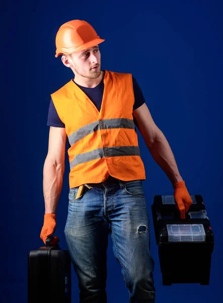 Trabalhador, faz-tudo, reparador, construtor no rosto calmo carrega sacos com ferramentas profissionais. Homem de capacete, chapéu duro segura caixa de ferramentas e mala com ferramentas, fundo azul. Conceito de reparador equipado — Fotografia de Stock