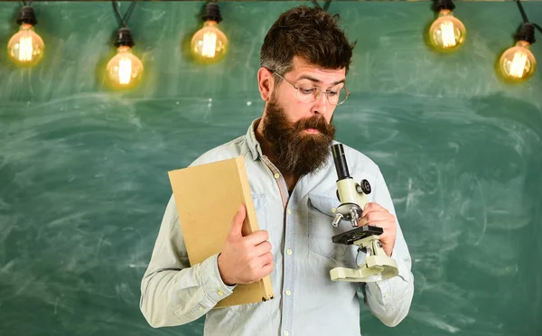 Mannen med skägg och mustasch på upptagen ansikte. Vetenskaplig forskning koncept. Lärare i glasögon håller boken och Mikroskop. Forskare håller boken och Mikroskop, svarta tavlan på bakgrund — Stockfoto