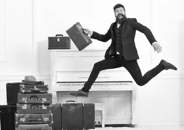 Macho attractive, elegant on cheerful face carries vintage suitcases, jumping. Long awaited vacation concept. Man with beard and mustache in suit carries luggage, luxury white interior background