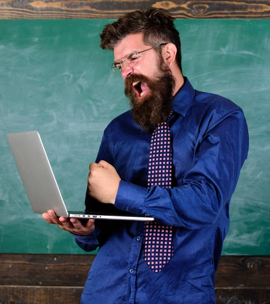 Technology blows his mind. Hipster teacher aggressive with laptop goes mad about modern technology. Can not get used to modern technology. Teacher bearded man with modern laptop chalkboard background — Stock Photo, Image