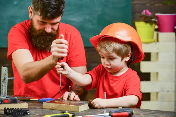 Trabajo en equipo en concepto de taller. Niño, niño ocupado en casco protector aprendiendo a usar destornillador con papá. Padre, padre con barba enseñando a su hijo a usar destornillador de herramientas — Foto de Stock