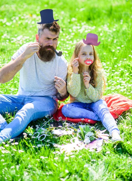 O pai e a filha sentam-se na relva no campo, fundo verde. Criança e pai posando com chapéu superior, lábios e atributos de cabine de fotos de tubulação. Conceito Aristocrates. Família passar o lazer ao ar livre — Fotografia de Stock