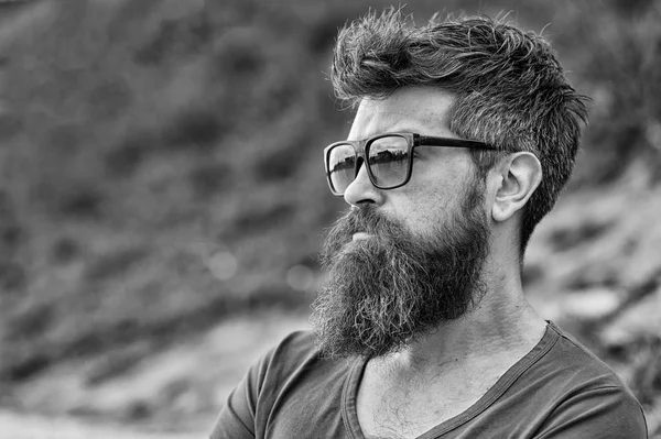 Hombre con barba y bigote en cara estricta, fondo natural, desenfocado. El hombre barbudo usa gafas de sol modernas. Hipster con barba se ve seguro mientras está de pie al aire libre. Barbería y concepto de estilo — Foto de Stock