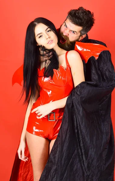 Vampire bites female neck. Vampire in cloak behind sexy devil girl. Couple in love play role game. Vampires victim concept. Man and woman dressed like vampire, demon, red background