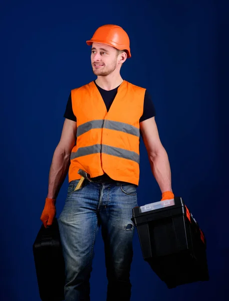 Handyman, reparador no rosto sorridente carrega sacos pesados com equipamento profissional. Homem de capacete, chapéu duro segura caixa de ferramentas e mala com ferramentas, fundo azul. Conceito de reparador profissional — Fotografia de Stock