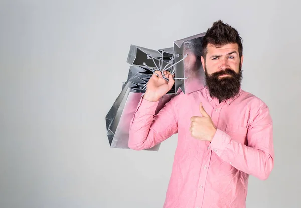 Kerl kauft ein und zeigt Daumen hoch. Hipster mit selbstbewusstem Gesicht empfiehlt zum Kauf. Black-Friday-Konzept. Mann mit Bart und Schnurrbart trägt Bündel Einkaufstüten, grauer Hintergrund — Stockfoto