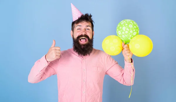 Hipster celebra cumpleaños, muestra gesto de pulgar hacia arriba. Concepto de niño de cumpleaños. El tipo con sombrero de fiesta con globos de aire celebra. Hombre con barba en la cara feliz celebrar globos de aire, fondo azul claro — Foto de Stock