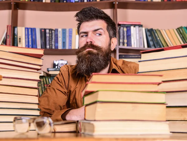 Homem no rosto estrito sentar entre pilhas de livros, enquanto estuda na biblioteca, estantes de livros no fundo. Professor ou estudante com barba sentar-se à mesa com óculos, desfocado. Conceito de bicho de biblioteca — Fotografia de Stock