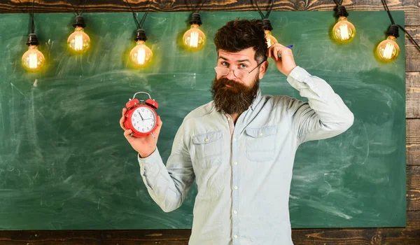 Hipster barbudo sostiene reloj, pizarra en el fondo, espacio de copia. Hombre con barba y bigote en cara olvidadiza con picazón en la cabeza. Profesor en anteojos sostiene despertador y pluma. Concepto de disciplina —  Fotos de Stock