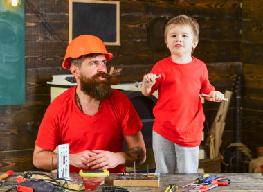 Evlat, çocuk neşeli cıvata veya vida, baba ile handcrafting sırasında eğlenmek tutar. Tamirci kavramı. Baba, ana okul atölyesinde farklı araçları kullanmak için kask öğretim oğlu sakallı