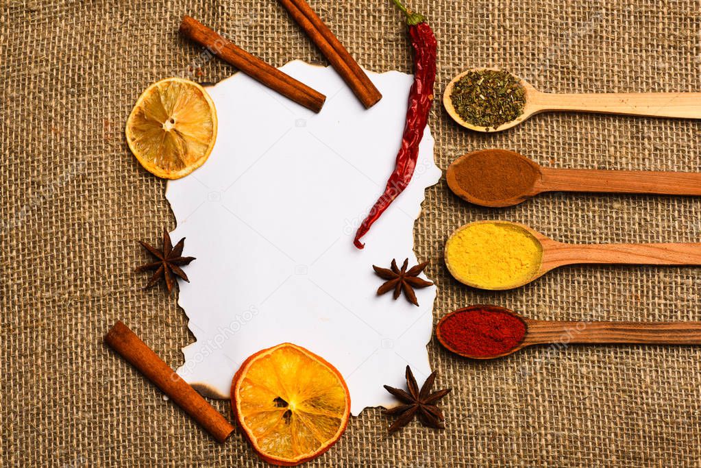 Piece of paper on sackcloth background. Cinnamon, dried orange and pepper, star anise around blank paper for recipe, copy space. Spoons with spices lay around white paper. Culinary recipe concept