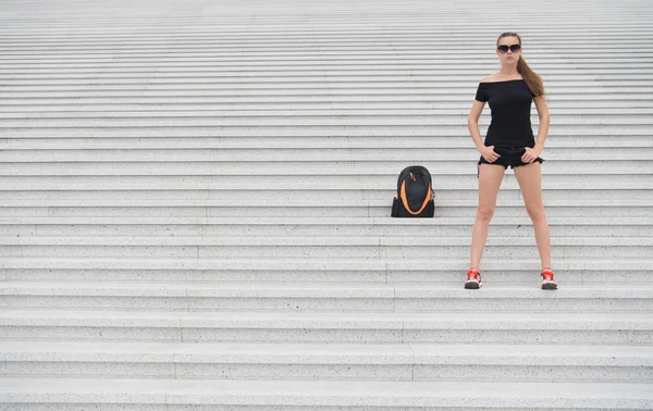 Μια τουρίστρια στέκεται με πέτρινες σκάλες κοντά στην τσάντα της. Έτοιμοι να εξερευνήσουμε νέα πόλη. Γυναικεία γυαλιά ηλίου μοντέρνα μαύρα ρούχα με τα πόδια Παρίσι. Διακοπές και ταξίδια έννοια. Τουριστικός οδηγός περιήγηση στα αξιοθέατα — Φωτογραφία Αρχείου