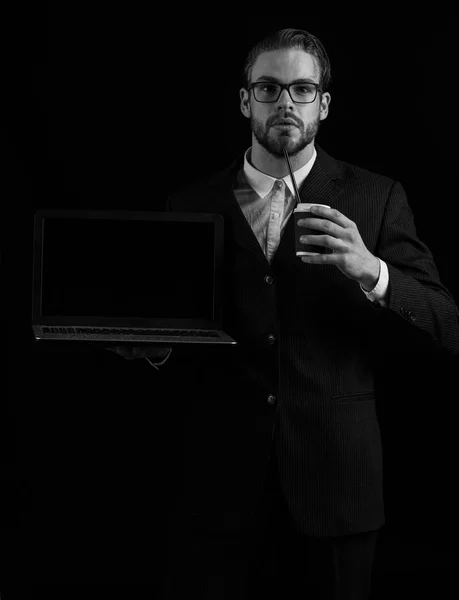 Bonito barbudo elegante homem de negócios em elegante terno preto em óculos segurando laptop e xícara de café em estúdio no fundo preto . — Fotografia de Stock