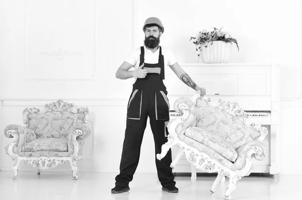 Relocating concept. Loader shows thumb up gesture. Courier delivers furniture in case of move out, relocation. Man with beard, worker in overalls and helmet lifts up armchair, white background