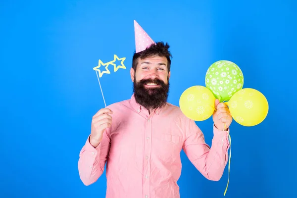 Homem com barba e bigode no rosto feliz segurar balões de ar, fundo azul. Conceito de aniversário. Hipster em óculos em forma de estrela celebra o aniversário. Cara em chapéu de festa com atributos de férias celebra — Fotografia de Stock