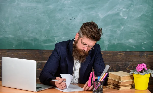 Перевірте домашнє завдання. Вчитель бородатий хіпстер з окулярами сидить на класному фоні. Вчитель сидить на столі з ноутбуком. Шкільний вчитель перевіряє домашнє завдання або тест. Паперова робота частина життя вчителів — стокове фото