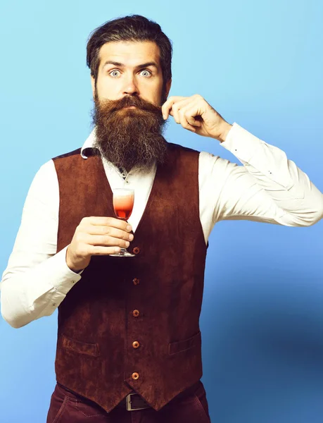 handsome bearded man with long beard and mustache on surprised face tasting glass of alcoholic shot in vintage suede leather waistcoat on blue studio background.