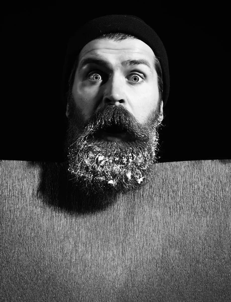handsome bearded man or guy in winter hat with fashionable mustache on surprised face and snow in beard near red paper on black background, copy space.
