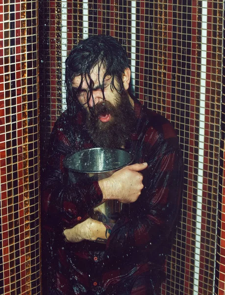 Homem bonito energizado, hipster barbudo com barba e bigode em camisa quadriculada vermelha molhada com água no chuveiro com balde de gelo de metal no fundo de mosaico colorido . — Fotografia de Stock