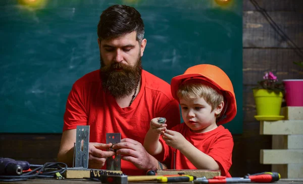 Home education concept. Father with beard and little son in classroom teaching to use tools, chalkboard on background. Boy, child in protective helmet makes by hand, repairing, does crafts with dad