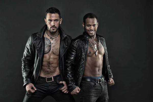 Models wearing dark jeans with heavy belts. Bikers in leather jackets on bare torsos. African and Hispanic men with sexy bodies. Male clothing, fashion concept. Brutal men on black background — Stock Photo, Image