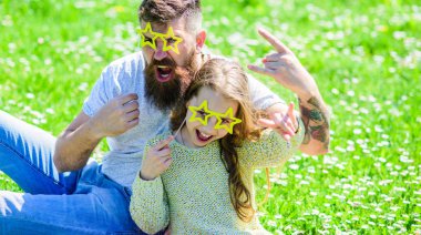 Rock star concept. Dad and daughter sits on grass at grassplot, green background. Child and father posing with star shaped eyeglases photo booth attribute at meadow. Family spend leisure outdoors clipart
