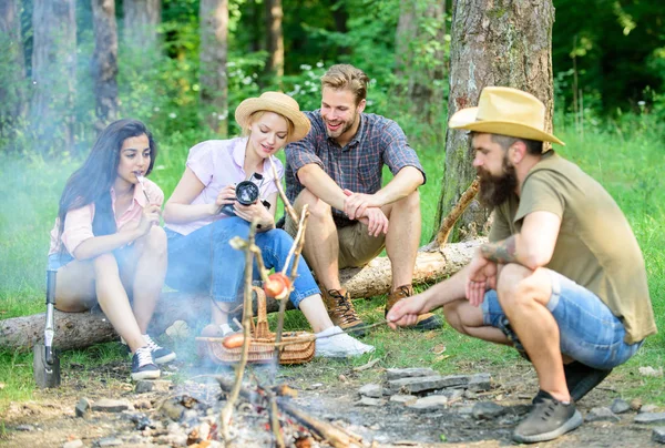 Friends group tourist relaxing near bonfire. Forest hike. Man roasting sausage while friends speak share impression and watching photos camera. Tourists having snack time with roasted over fire food