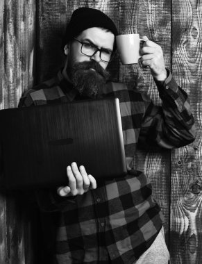 Sakallı, beyaz tenli hippi elinde bardakla dizüstü bilgisayar tutuyor.