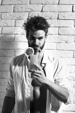 Genç, sakallı, seksi, maço, şık sakallı, düğmesiz beyaz gömlekli, kaslı, çıplak vücutlu, beyaz tuğla duvar arkasında alkollü, turuncu bir kokteyl bardağı tutan adam.