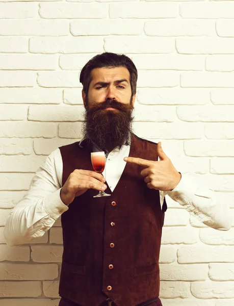 handsome bearded man with long beard and mustache has stylish hair on funny face holding glass of alcoholic shot in vintage suede leather waistcoat on white brick wall studio background