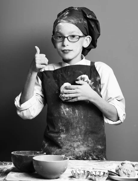 Young boy cute cook chef in uniform and hat on stained face flour with glasses standing near table with colorful bawls and holding tasty chocolate cakes on red studio background — Stock Photo, Image