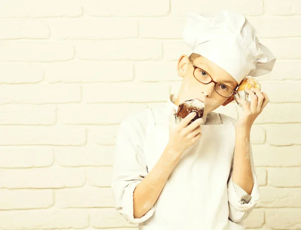 young boy cute cook chef in white uniform and hat on stained face flour with glasses holding chocolate cakes on brick wall background, copy space.