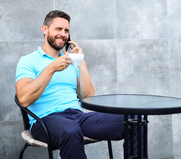 Time to relax. Brief coffee break provides employees with quick ways to relax. Coffee break brings physical and mental wellbeing. Man sit terrace and drink cappuccino speak phone grey wall background