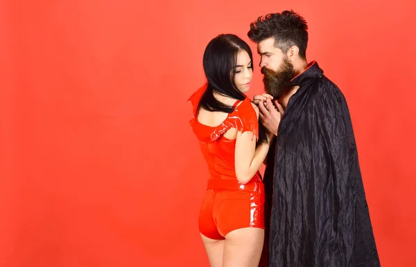 Couple in love, perfect match. Vampire in cloak and sexy devil girl holds hands. Couple on pensive faces play role game. Devil love concept. Man and woman dressed like vampire, demon, red background