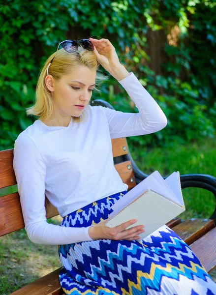 书是她的爱好。女金发女郎在公园看书时休息放松。女孩坐板凳放松与书, 绿色自然背景。阅读文学作为爱好。喜欢看书的女孩 — 图库照片