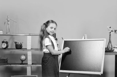 Kız boş yazı tahtası üzerinde kopya alanı yazar. Eğitim ve okul kavramı dön. Sarı duvar arka plan üzerinde çocuk ve okul malzemeleri. Emin yüz ve ponytails ile kız öğrenci sınıfta standları