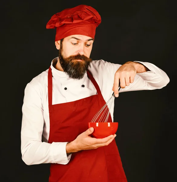 Kock med nyfikna ansikte i vinrött uniform innehar skål, visp — Stockfoto