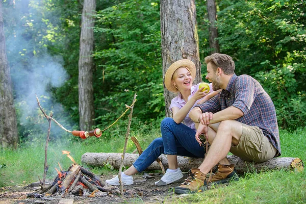 Have snack. Couple relaxing sit on log having snacks. Family enjoy romantic weekend in nature. Girl offers eat apple while they waiting roasted food. Picnic roasting food over fire. Cooking at picnic