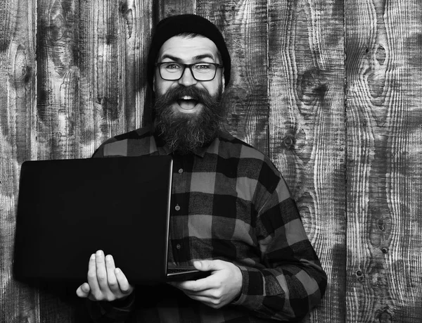 Homme barbu, longue barbe. Brutal caucasien souriant heureux hipster non rasé tenant ordinateur portable en rouge noir chemise à carreaux avec chapeau et lunettes sur fond de studio en bois vintage brun. — Photo
