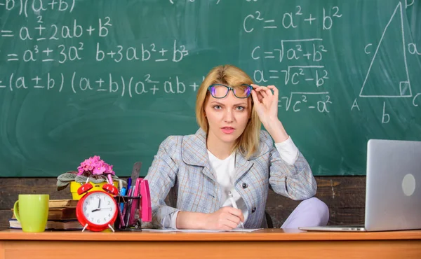 Tiene dudas en su informe o conocimiento. El profesor se asomaba sospechosamente por las gafas. Profesor mujer sentarse mesa aula pizarra fondo. Maestro cara dudosa escucharte — Foto de Stock