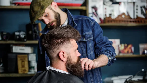 Barbiere occupato con governare la barba del cliente hipster, forniture di bellezza sullo sfondo. Il concetto di toelettatura. Hipster con barba ricoperta di mantello che serve da barbiere professionista in elegante barbiere — Foto Stock