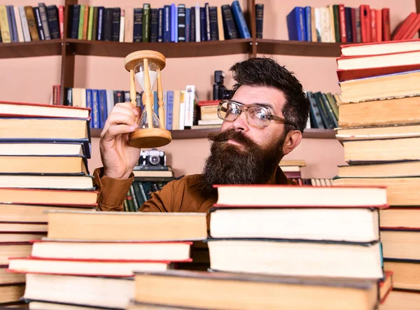 Mann mit nachdenklichem Gesicht hält Sanduhr während des Studiums, Bücherregale im Hintergrund. Lehrer oder Schüler mit Bart studieren in der Bibliothek. Mann, Wissenschaftler mit Brille blickt auf Sanduhr. Zeitflusskonzept — Stockfoto