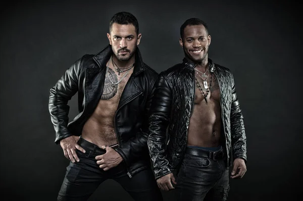 Dos modelos masculinos posando juntos. Concepto de deseo y seducción. Un africano con sonrisa alegre. Hombre hispano con tatuaje geométrico en el pecho peludo. Culturistas con chaquetas de cuero en torsos desnudos — Foto de Stock