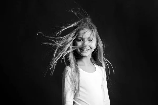 Salón de belleza y peluquería. Modelo de niño sonriendo con el pelo largo que sopla. Chica con sonrisa adorable sobre fondo oscuro. Moda, mira, peinado. Concepto de infancia feliz — Foto de Stock