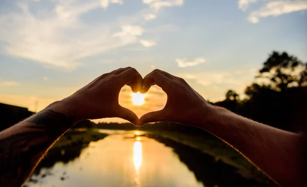 Gesto do coração em frente ao pôr do sol acima da superfície da água do rio, desfocado. Os melhores lugares para encontros românticos. Mãos masculinas em forma de coração gesto símbolo de amor e romance. Pôr do sol atmosfera romântica — Fotografia de Stock