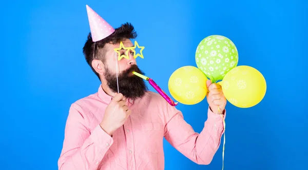 Mann mit Bart im Gesicht hält Luftballons, blauer Hintergrund. Hipster in sternförmiger Brille bläst ins Partyhorn. Mann im Partyhut mit Feiertagsattributen feiert. Überraschungskonzept — Stockfoto