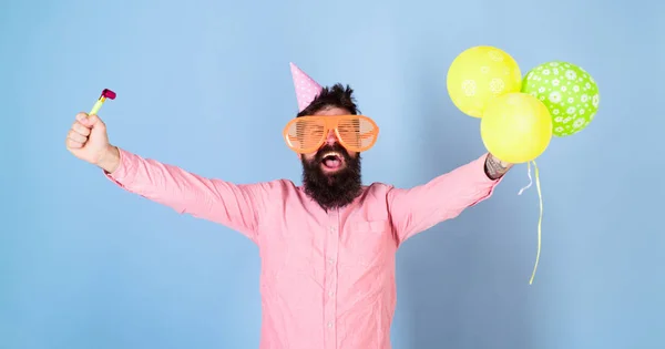 Homem com barba e bigode no rosto feliz detém balões de ar, fundo azul claro. Conceito de festa. O tipo de chapéu de festa com atributos de férias celebra. Hipster em óculos gigantes celebrando aniversário — Fotografia de Stock