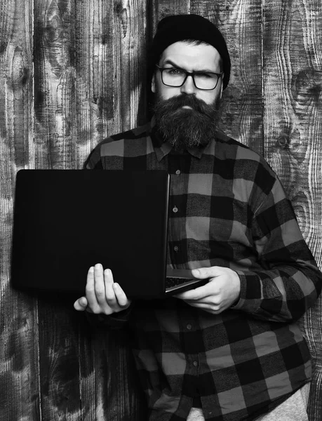 Homem barbudo, barba comprida. Brutal caucasiano sério sem barba hipster segurando laptop em vermelho camisa xadrez preto com chapéu e óculos em marrom vintage estúdio de madeira fundo. — Fotografia de Stock