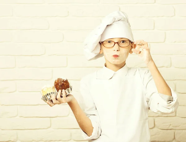 Mladý chlapec roztomilý kuchař kuchař v bílé uniformě a klobouk na skvrnité tvář mouky s brýlemi drží čokoládové dorty na cihlové zdi pozadí, kopírovat prostor. — Stock fotografie
